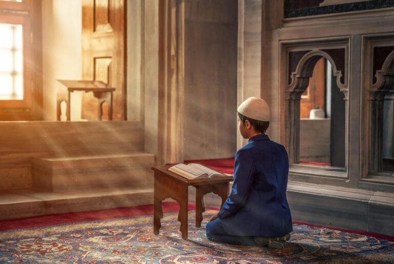 About A young boy kneels in prayer inside a mosque with sunlight streaming through the window.