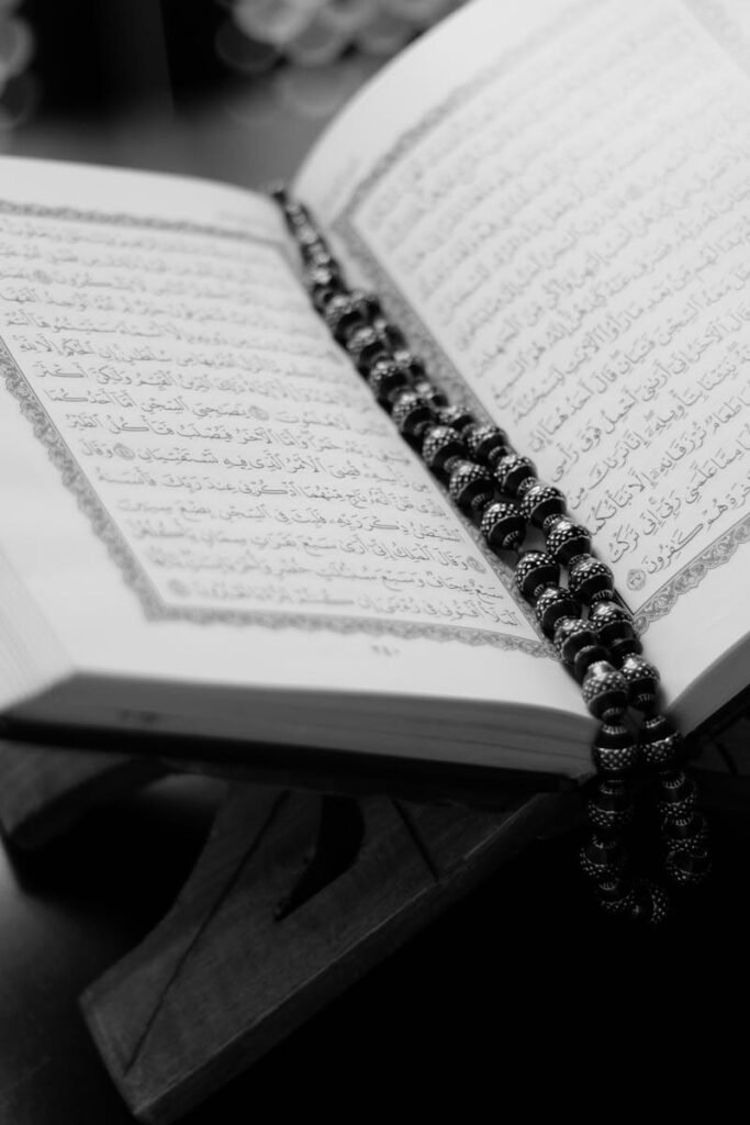 About A black and white close-up of an open Quran on a wooden stand with prayer beads, symbolizing Islamic faith and devotion.